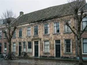 Verweerde bakstenen rijtjeswoning met enkele beglazing, brokkelig voegwerk en verouderd pannendak op bewolkte Nederlandse straat in de winter.