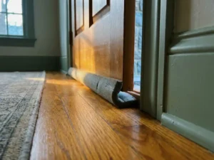Tochtstrip op drempelplaat drukt schuim afdichting tegen gepolijste houten vloer, warm amber licht gloeit door deurkier.